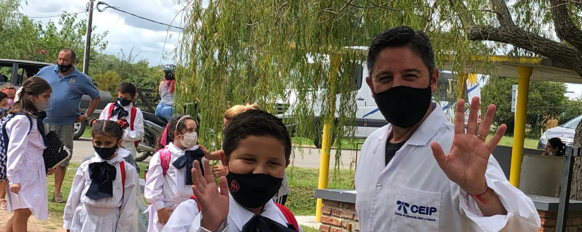 Darío Greni junto a un alumno de la escuela. Darío Greni y un alumno de la escuela, vestidos con túnica, saludan mirando a la cámara. Detrás se ven otros estudiantes. Se encuentran fuera de la escuela.