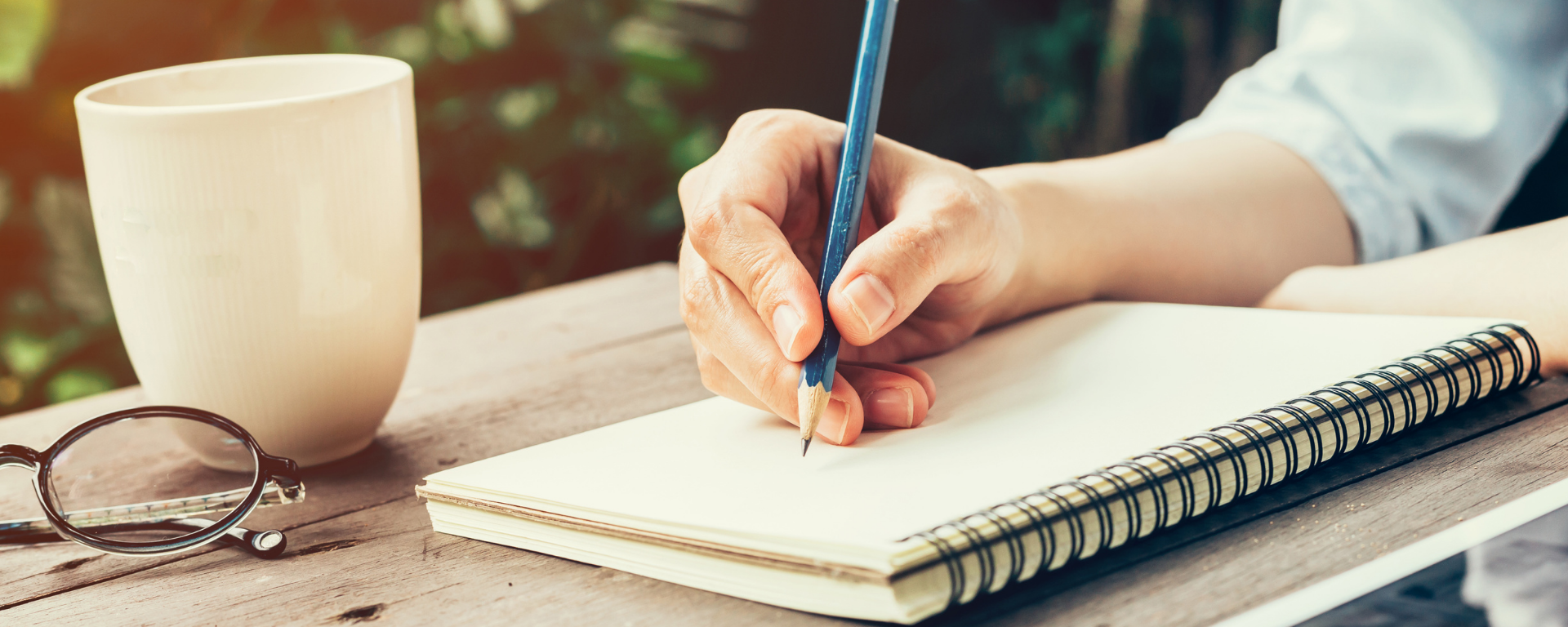 Taller de escritura: mano escribiendo con lápiz sobre cuaderno, en una mesa en exteriores, con una taza de café